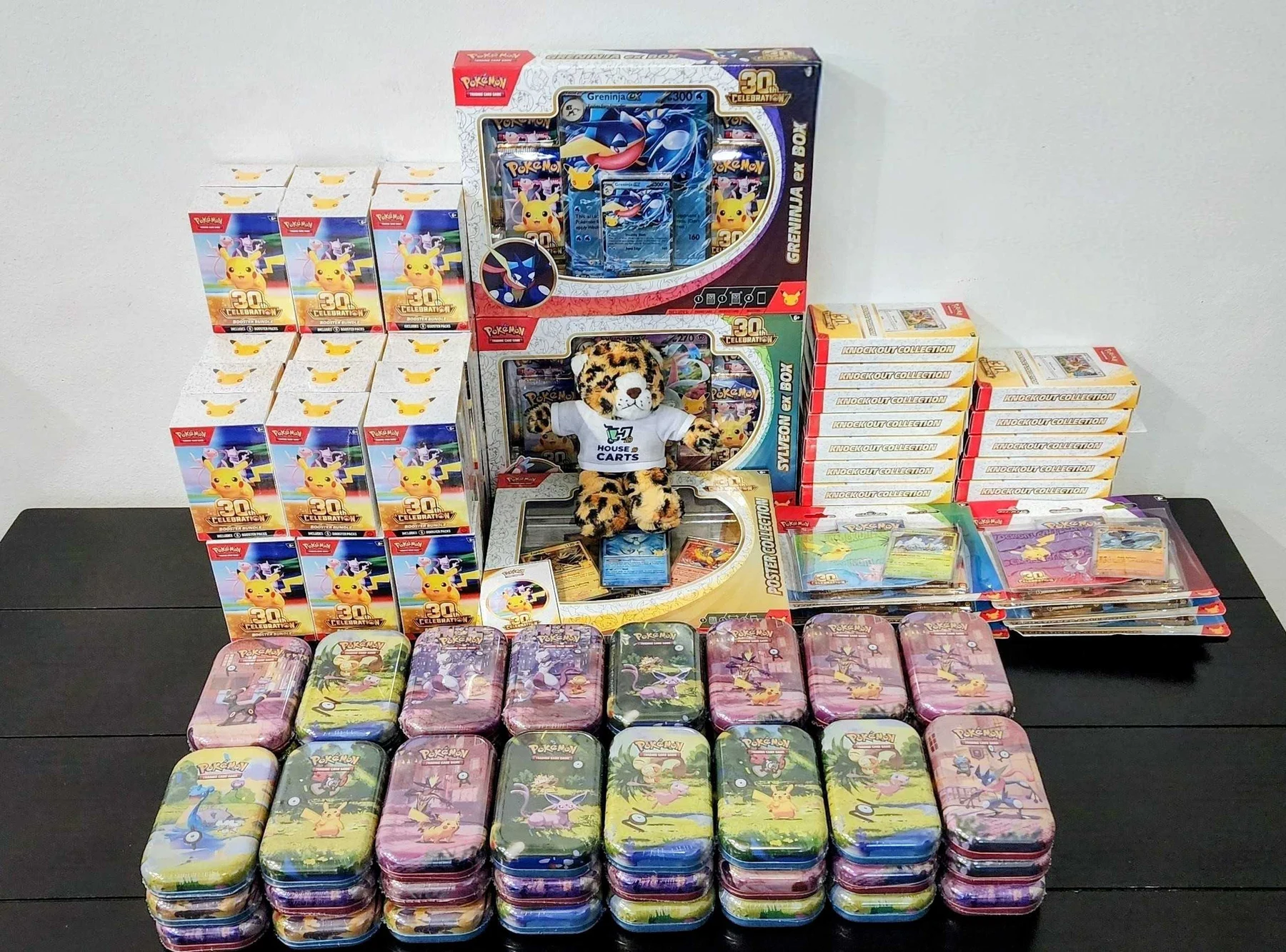 Member photo of a tabletop filled with Pokémon tins, 30th Celebration Booster Bundles, collection boxes and an HOC mascot.